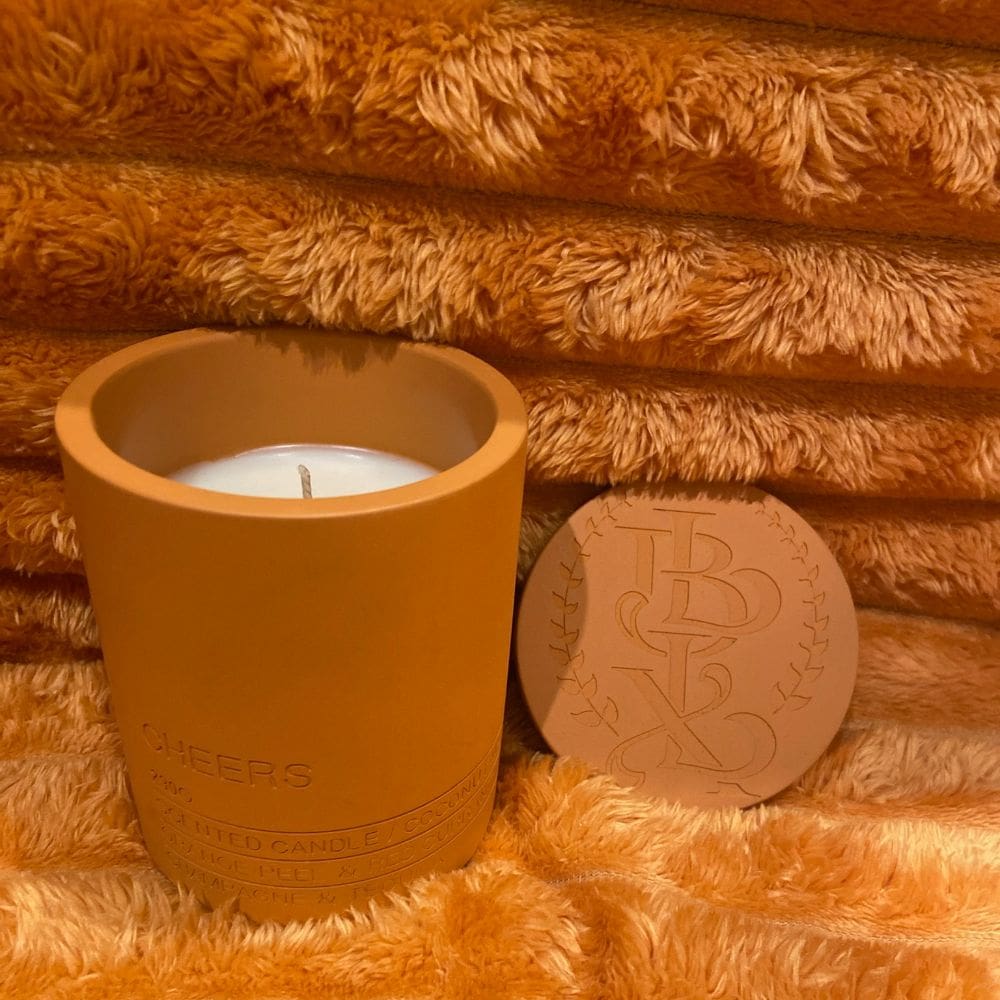 A lit candle in a brown holder with a matching round lid on a textured brown surface.
